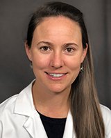 Headshot of a young female physician. Dr. Anderson has long straight brown hair. Dr. Anderson is wearing a black crew shirt under white coat. She is sitting in front of a light grey background.