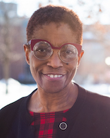 Headshot of UVMMC Board Trustee, Noma Anderson, standing outside the medical center. She is wearing a red and black check blouse and a black cardigan. She is wearing red glasses.