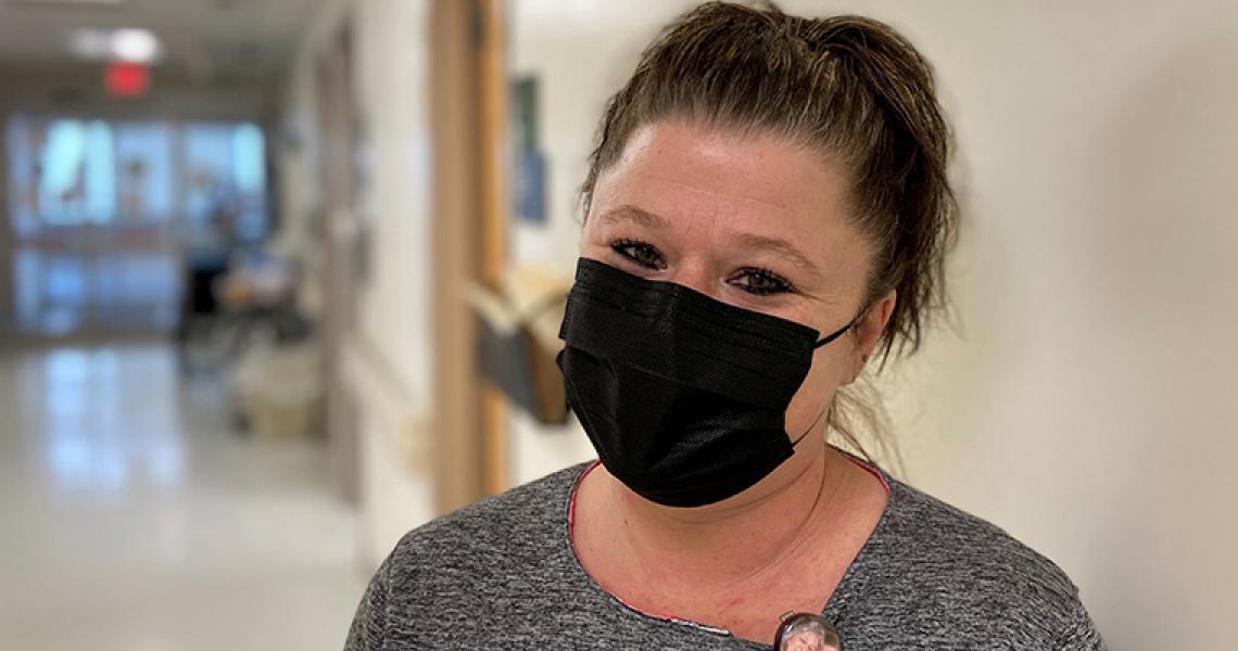 Headshot of Melissa Stoddard, a nurse wearing a black face mask. Melissa is a middle aged woman, has brown hair pulled into a pony tail and she is wearing a grey shirt. She is standing in the hospital hallway, facing the camera, and smiling. 