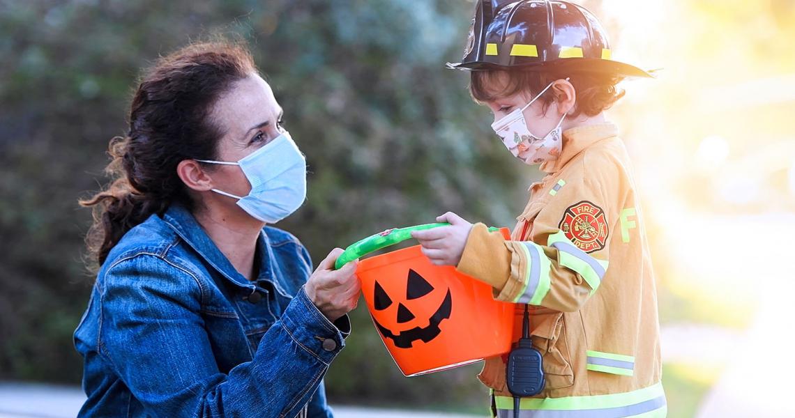 Masked woman giving Halloween candy to child wearing a firefighter costume and a mask