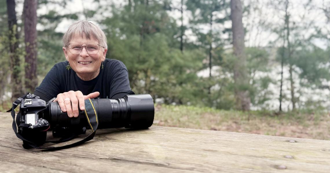 Portrait Raven Schwan-Noble sitting at a picnic bench with one hand on her Camera.