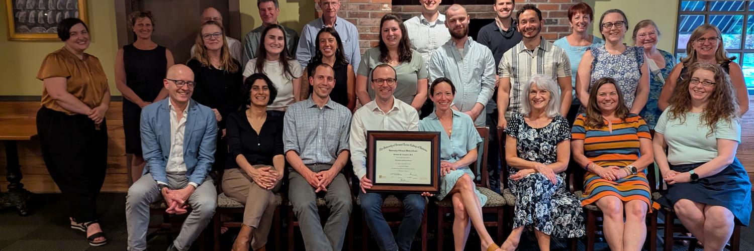 Team of staff and faculty sitting in front of a fireplace. The person in the front is holding a diploma.