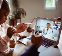 Father holding thermometer with son on lap talking with doctor on laptop