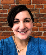 Photo of Christa Hagan-Howe smiling, wearing a blue shirt and in front of a brick wall