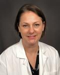Headshot of a young female physician. She has short brown hair. She is wearing a black blouse under a white coat. She is sitting in front of grey backdrop.