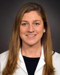 Headshot of a young physician. She has long brown hair. She is wearing a black crew neck shirt under a white coat.