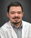 Headshot of a middle-aged male provider. He has dark brown hair and close shaved-beard. He is wearing a light grey collared shirt under a white coat.