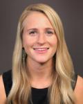 Headshot of a young female physician. Hillary Stevens has long blond hair and is wearing a black sleevless crew-neck blouse. She is sitting in front of a light grey background.