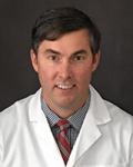 Headshot of a middle-aged male vascular surgeon. He has short brown hair and clean-shaven. He is wearing a collared shirt and red striped tie under his doctor's white coat. He is sitting against a grey background.