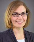Headshot of a middle aged physician. She is wearing black glasses. She is sitting in front of a light grey background and wearing a white crew neck top under a dark grey sweater.