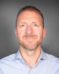 Headshot of a male nurse practitioner. He is posing in front of a grey background and wearing a light blue collared shirt.
